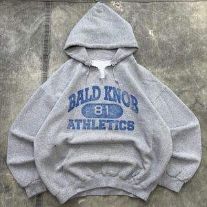 Puede incluir: Una sudadera con capucha de color gris jaspeado que presenta un gráfico azul desgastado con el texto BALD KNOB 81 ATHLETICS. La prenda tiene un cuello sin rematar con un pequeño corte en V y un bolsillo tipo canguro en la parte frontal.