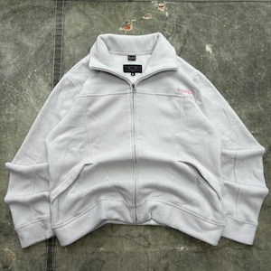 May include: A light gray fleece jacket with a full zipper closure and a high collar. The jacket features a small logo on the chest and a brand label near the collar. The jacket has two front pockets and is made of a soft, warm material.