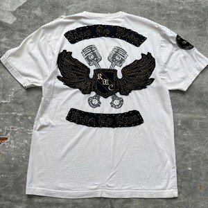 May include: White t-shirt with a black and gold embroidered patch featuring a pair of crossed pistons with wings and a shield with the letters "RMC". The patch is surrounded by a black studded banner with the text "Ride To Live".
