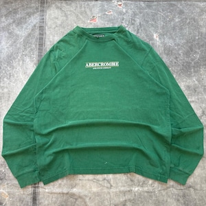 May include: A long-sleeve green crewneck shirt featuring the text ABERCROMBIE AND FITCH COMPANY printed in white on the chest. The garment has a ribbed neckline and raglan sleeves, with a simple, solid-coloured design throughout the fabric.