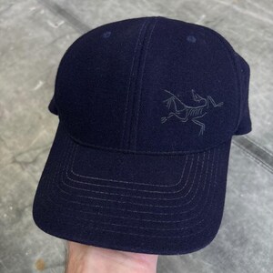 May include: A dark navy blue baseball cap featuring a structured crown and a curved brim with tonal stitching. The front panel displays a dark grey embroidered logo of a skeletal creature, positioned slightly to the right of the center seam.
