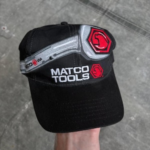 Può includere: Cappellino da baseball nero con le parole "MATCO TOOLS" ricamate in bianco. Il cappellino presenta un design a chiave inglese argentato e rosso e la scritta "USA". Il cappellino è realizzato in un tessuto resistente.