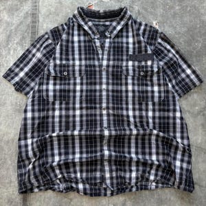 Może przedstawiać: Koszula z krótkim rękawem w czarno-białą kratę, z dwiema kieszeniami na piersi i zapięciem na guziki. Koszula ma klasyczny kołnierzyk i naszywkę na lewej piersi. Tkanina ma miękki, zużyty wygląd.