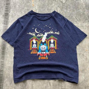 May include: Dark blue t-shirt with a colourful graphic of Thomas the Tank Engine. The design includes a crescent moon, stars, and train carriages. The shirt has a crew neck and short sleeves.