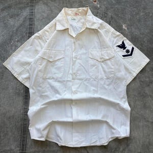 Puede incluir: Camisa de manga corta color crema con dos bolsillos en el pecho y cierre de botones. Un parche azul marino con un águila y una estrella está en la manga izquierda. La camisa tiene un cuello clásico y está hecha de tela ligera.