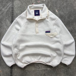 May include: A cream-colored long-sleeve fleece pullover featuring a snap-button placket, a high collar, and a single chest pocket with a flap. The garment includes a dark blue Gap Arctic Light Fleece label at the neck and a small rectangular logo patch.