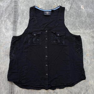 Może przedstawiać: Czarny top bez rękawów z dwiema przednimi kieszeniami i dekoracyjnymi panelami z koronki po bokach. Top ma zapięcie na guziki z przodu i małe logo w pobliżu dekoltu. Top jest wykonany z miękkiej, lekkiej tkaniny.