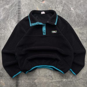 May include: Black fleece pullover with a teal collar, placket, and trim. The pullover has a button closure and a small logo patch on the chest. A cozy and warm garment for outdoor activities.
