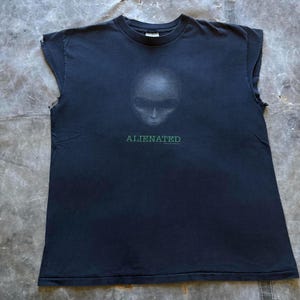 May include: Black sleeveless t-shirt featuring a gray alien face graphic and the word "ALIENATED" in green. The shirt has a crew neck and a relaxed fit, perfect for casual wear. The fabric appears to be slightly worn, giving it a vintage look.