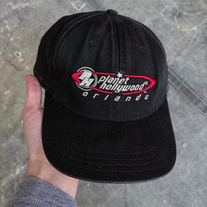 Può includere: Cappellino da baseball nero con il logo Planet Hollywood Orlando ricamato in rosso, bianco e nero. Il cappellino ha una visiera curva e una corona strutturata. Il testo "Planet Hollywood Orlando" è arcuato sopra la visiera.