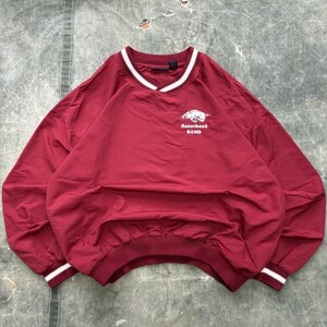 May include: A maroon pullover windbreaker with a V-neck collar and elastic cuffs. The collar and cuffs have white stripes. The text "Razorback BAND" is printed in white, along with a graphic of a hog.