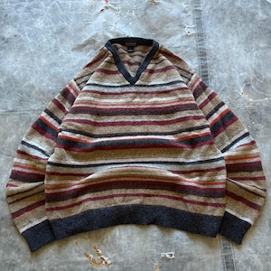 May include: A V-neck sweater with horizontal stripes in various colours. The stripes include shades of brown, red, and grey. The neckline, cuffs, and hem are a darker grey. The sweater is made from a soft, textured fabric.