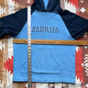 May include: A light blue hooded sweatshirt with navy blue sleeves. The sweatshirt has a large "CAROLINA" graphic on the front.