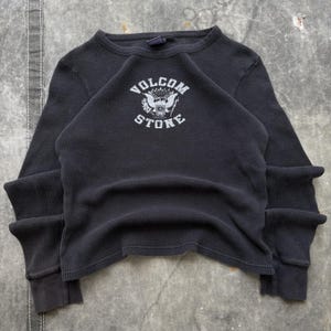 May include: Dark gray waffle-knit long-sleeve shirt with a crew neck and ribbed cuffs. The shirt features a white graphic on the chest with the text "VOLCOM STONE" and a stylized emblem. The shirt is laid flat on a textured surface.