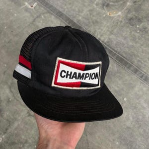 May include: Black Champion baseball cap with a mesh back and a curved brim. The cap features a rectangular patch with the word "CHAMPION" in white letters on a red and white background. Red and white stripes are on the side.