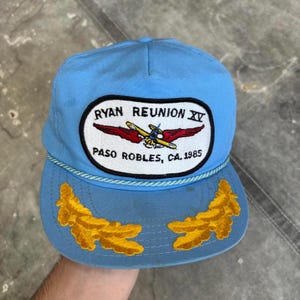 May include: Pale blue baseball cap with a curved peak. The cap has an embroidered patch with the text "RYAN REUNION XV" and "PASO ROBLES, CA. 1985" and an airplane graphic. The peak has two embroidered gold leaf designs.
