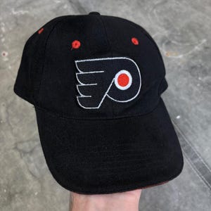 May include: A black baseball cap featuring an embroidered Philadelphia Flyers logo on the front. The logo consists of a stylised black letter P with orange wings and a central orange circle. The cap includes small red ventilation eyelets on top.