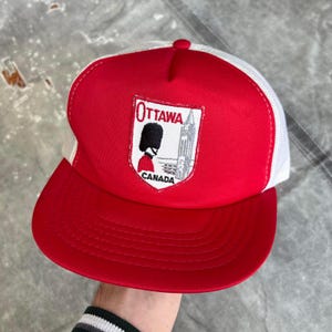 Puede incluir: Gorra de camionero roja y blanca con visera y corona rojas, y parte trasera de malla blanca. La parte delantera presenta un parche con la palabra "Ottawa", una ilustración de un guardia y la palabra "Canadá".
