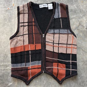May include: A sleeveless knit vest featuring a full-length front zipper and a V-neckline. The garment displays a patchwork pattern of black, brown, tan, orange, and gray rectangular blocks with thin white and orange grid lines. A White Stag label is visible.