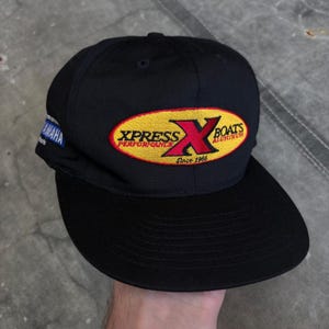 Könnte beinhalten: Eine schwarze Baseballkappe mit einem gestickten gelben ovalen Aufnäher auf der Vorderseite, der den roten Schriftzug XPRESS PERFORMANCE X BOATS ALUMINUM Since 1966 trägt. Ein kleineres blau-weißes Yamaha-Logo ist seitlich aufgestickt.