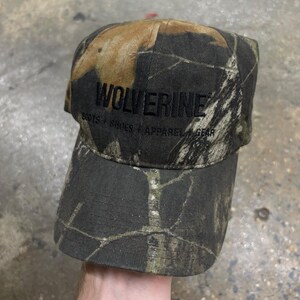 May include: A camouflage baseball cap featuring a brown, green, and black leaf pattern. The front of the cap displays the black embroidered text WOLVERINE with the smaller text BOOTS + SHOES + APPAREL + GEAR printed directly underneath it.
