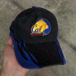 May include: Black baseball cap with a blue brim and accents. The cap features an embroidered logo with a yellow bear and the word "Cal" above "Aquatics" in red. The cap is made of a durable fabric.
