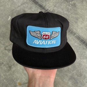 May include: Black baseball cap with a flat brim. The cap features a rectangular patch with a light blue background, the text "Phillips 66 AVIATION", and a winged emblem in white and red.