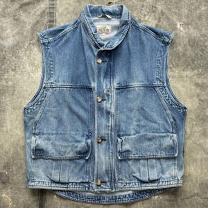 Puede incluir: Un chaleco vaquero azul claro sin mangas. Presenta un cuello, cuatro bolsillos delanteros y cierres de botón. El nombre de la marca "Redhead" es visible en el cuello. El chaleco está hecho de mezclilla resistente.