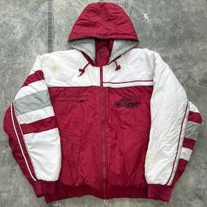 Op de afbeelding: Een hooded jack in bordeauxrood, wit en grijs. De jas heeft een bordeauxrode body, witte schouders en mouwen, geaccentueerd door grijze strepen. Een logo is op de borst geborduurd. De jas heeft een capuchon met trekkoord en geribde manchetten.