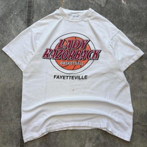 May include: White t-shirt featuring the words "LADY RAZORBACK BASKETBALL" in red and black lettering over an orange basketball graphic. The word "FAYETTEVILLE" is printed below the graphic.