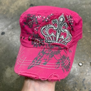 Puede incluir: Una gorra de estilo militar de color rosa brillante con visera desgastada y deshilachada. La gorra está decorada con un estampado floral gris, pedrería plateada y un parche central bordado de una corona con costuras plateadas y bordes deshilachados.