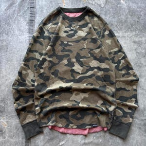 May include: Camouflage-patterned long-sleeve shirt in shades of green, brown, and black. The shirt has a waffle knit texture and a crew neck. The cuffs and neckline are dark gray. The inside of the shirt is pink.