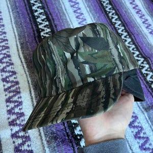 Puede incluir: Una gorra de béisbol con estampado de camuflaje y una solapa de oreja negra. La gorra tiene un estampado de camuflaje marrón, verde y negro.