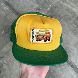 Op de afbeelding: Een gele en groene truckerpet met een patch met een rode en gele bus en het woord "CHEVY". De pet heeft een groene mesh achterkant en een groene klep. De voorkant van de pet is geel.
