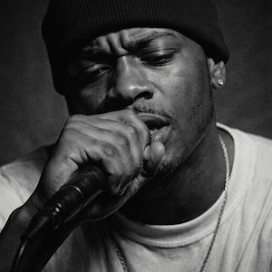 May include: Black and white portrait of a person wearing a black beanie and a white t-shirt, holding a microphone. A silver chain necklace is visible. The person's expression is focused, with their mouth near the microphone.