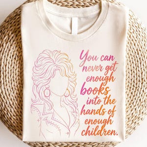 May include: A cream-colored t-shirt featuring a pink and orange gradient line art illustration of a person with voluminous hair and the text: You can never get enough books into the hands of enough children. The design is centered on the front.