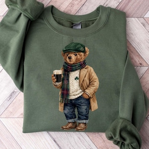May include: Green sweatshirt featuring a graphic of a bear wearing a green cap, plaid scarf, tan coat, and jeans, holding a dark beverage. The bear has a shamrock on its sweater. The sweatshirt is made of a soft material and has long sleeves.