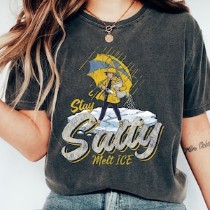 May include: Dark gray t-shirt featuring a cartoon illustration of a person in a yellow raincoat and umbrella, standing on ice. The text "Stay Salty Melt ICE" is in a stylized font, with yellow and white accents.