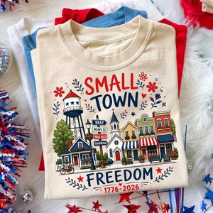 May include: A stack of folded t-shirts in red, white, and blue. The top shirt is beige with a colorful graphic that reads "SMALL TOWN FREEDOM 1776-2026" featuring a water tower, buildings, and floral accents. Decorative elements include disco balls and star-shaped confetti.