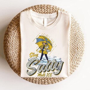 May include: Cream-colored t-shirt featuring a graphic of a person in a yellow raincoat and umbrella standing on ice. The text "Stay Savvy Melt ICE" is below the illustration. The shirt is laid on a woven, circular surface.