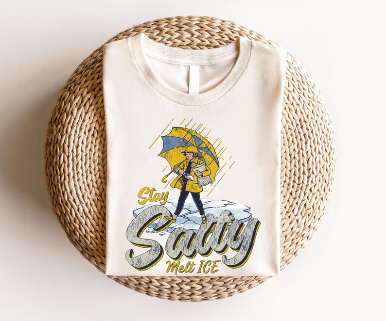 May include: Cream-colored t-shirt featuring a graphic of a person in a yellow raincoat and umbrella standing on ice. The text "Stay Savvy Melt ICE" is below the illustration. The shirt is laid on a woven, circular surface.