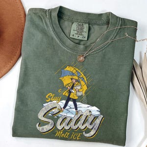 May include: Olive green t-shirt with a graphic of a person under a yellow and blue umbrella, standing on ice. The text on the shirt reads "Stay Salty Melt ICE". A silver necklace and a brown hat are also visible.