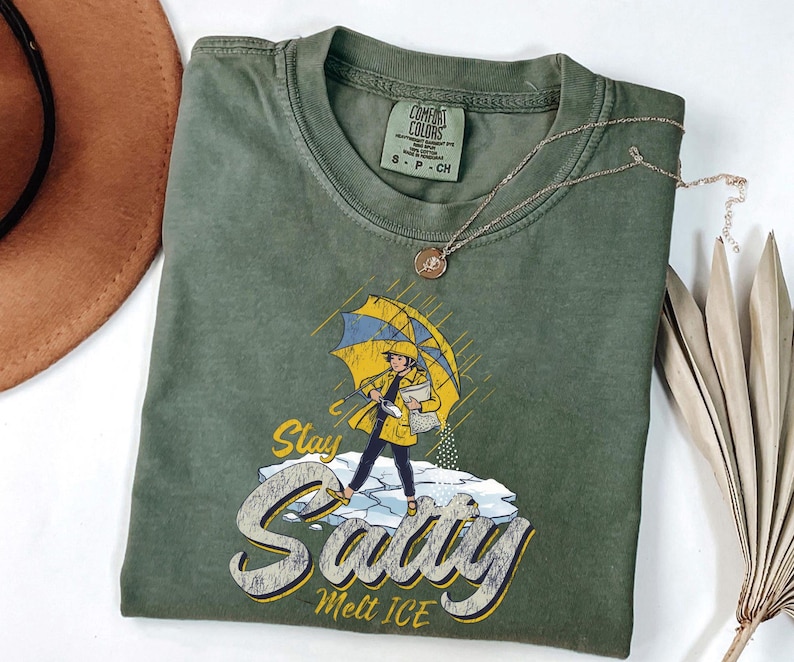 May include: Olive green t-shirt with a graphic of a person under a yellow and blue umbrella, standing on ice. The text on the shirt reads "Stay Salty Melt ICE". A silver necklace and a brown hat are also visible.