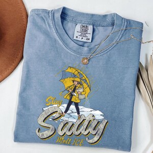 May include: A light blue Comfort Colors t-shirt featuring a graphic of a person in a yellow raincoat and umbrella, standing on ice. The text "Stay Salty Melt Ice" is below the graphic. A gold necklace is draped across the shirt.
