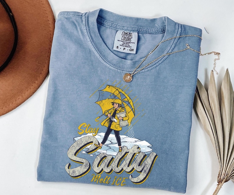 May include: A light blue Comfort Colors t-shirt featuring a graphic of a person in a yellow raincoat and umbrella, standing on ice. The text "Stay Salty Melt Ice" is below the graphic. A gold necklace is draped across the shirt.