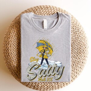 May include: A heather gray t-shirt featuring a graphic of a person in a yellow raincoat and umbrella standing on ice, with the words "Stay Savvy Melt Ice" in a stylized font. The shirt has a crew neck.