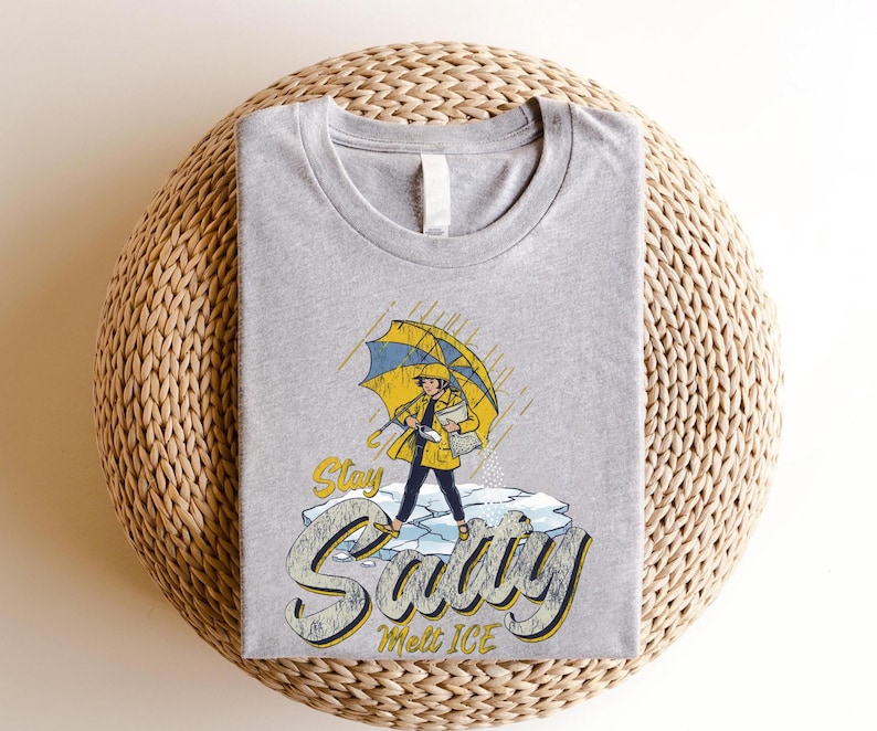 May include: A heather gray t-shirt featuring a graphic of a person in a yellow raincoat and umbrella standing on ice, with the words "Stay Savvy Melt Ice" in a stylized font. The shirt has a crew neck.