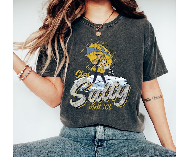 May include: Dark gray t-shirt featuring a cartoon illustration of a person in a yellow raincoat and umbrella, standing on ice. The text "Stay Salty Melt ICE" is in a stylized font, with yellow and white accents.