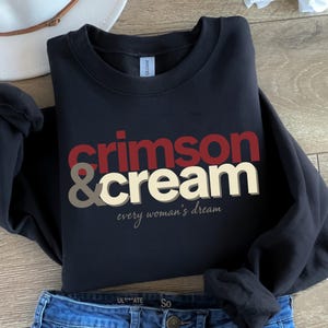 May include: A black crewneck sweatshirt with the words "Crimson & Cream" in red, cream, and taupe lettering. The phrase "every woman's dream" is printed below the main text in a script font. The sweatshirt is paired with blue jeans.