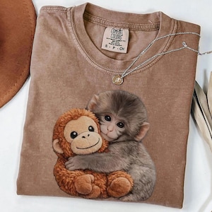 May include: A brown Comfort Colors t-shirt featuring a graphic of two monkeys hugging. The shirt has a crew neck and short sleeves. A silver necklace with a small pendant is draped across the shirt.